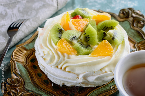 Gentle, light, airy, delicious pavlova dessert with slices of orange and kiwi on top in a nice vintage wooden tray next to dessert fork on a linen napkin and cup of tea, closeup