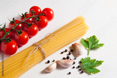 Bunch of raw spaghetti, cherry tomatoes, peppers, garlic cloves and parsley leaves on a white wooden background.