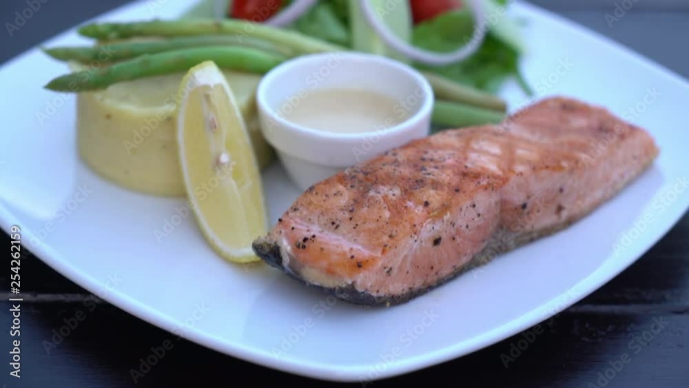 A close up shot of a white squared plate with fried salmon, sliced lemon, and some vegetables on it. A close up shot of a white squared plate with fried salmon, sliced lemon, and some vegetables on it.