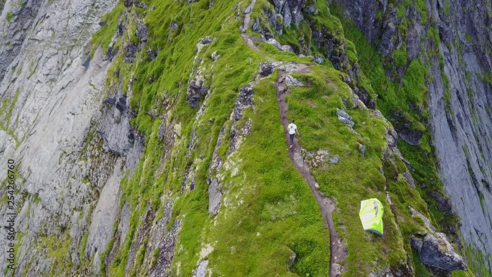 Young man, facing away, hiking on narrow footpath on top of mountain ridge, walking past a yellow tent, high angle aerial follow shot, outdoor adventure and sports in Scandinavia, Reinebringen Young man, facing away, hiking on narrow footpath on top of mountain ridge, walking past a yellow tent, high angle aerial follow shot, outdoor adventure and sports in Scandinavia, Reinebringen