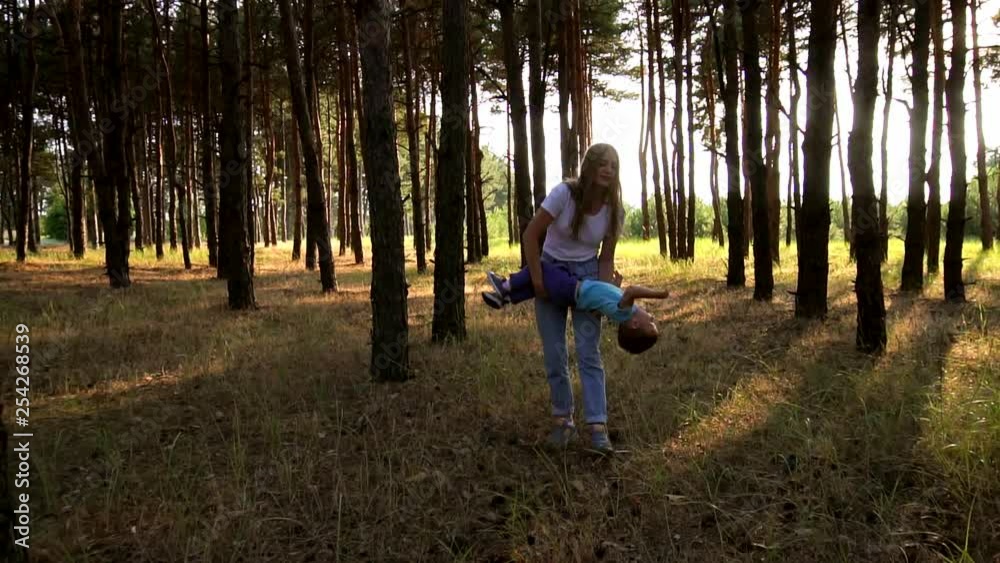 Young mother playing with adorable little fid on nice evening walk through evergreen forest filled with sunset sun rays