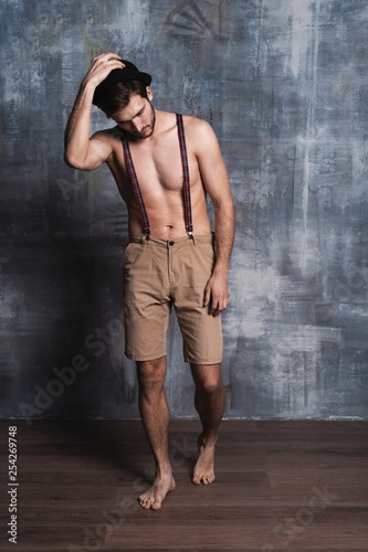 Handsome guy fashion sexy young bearded macho man model with suspenders on pants on gray background. © opolja