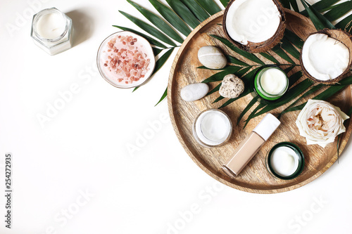 Styled beauty composition. Skin creams, makeup bottle, rose and pebble stones on wooden tray. Coconuts, tropical palm leaves decoration. Cosmetics, spa concept. Empty space, flat lay, top view.