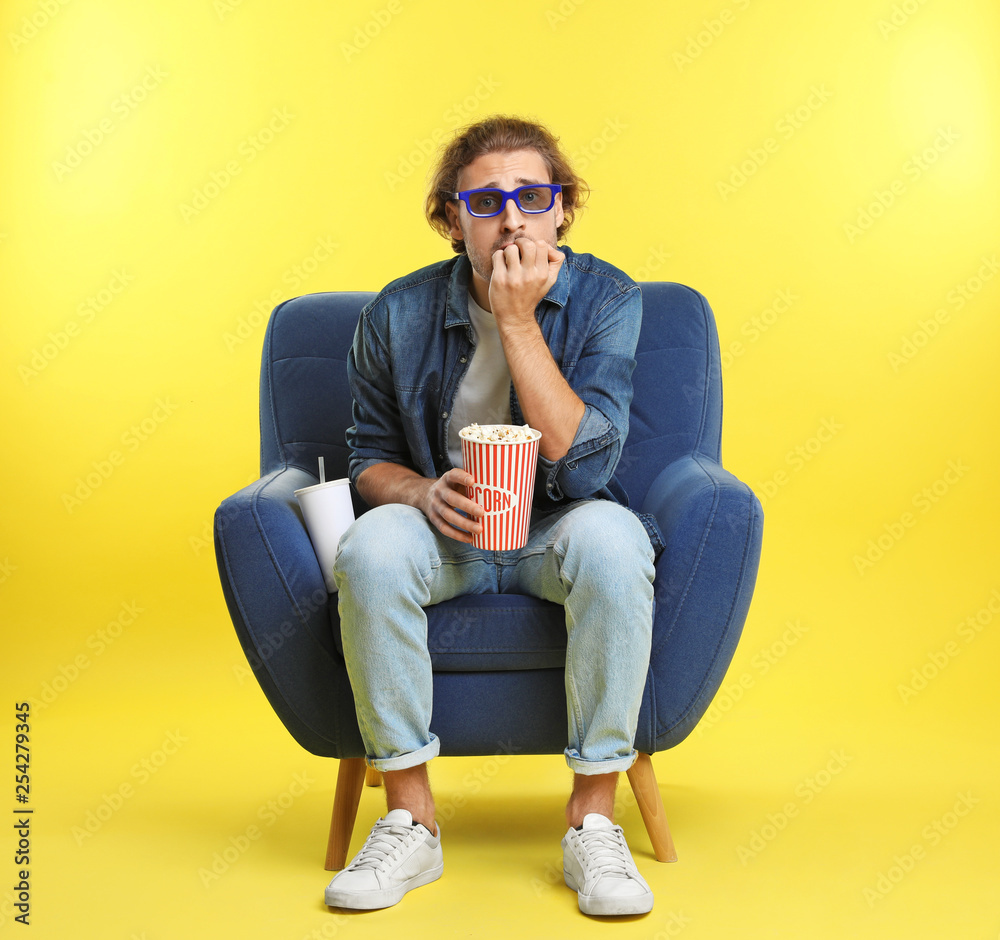 Emotional man with 3D glasses, popcorn and beverage sitting in armchair during cinema show on color background Emotional man with 3D glasses, popcorn and beverage sitting in armchair during cinema show on color background
