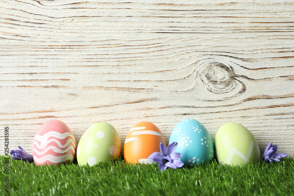 Colorful painted Easter eggs with flowers on green grass against wooden background, space for text