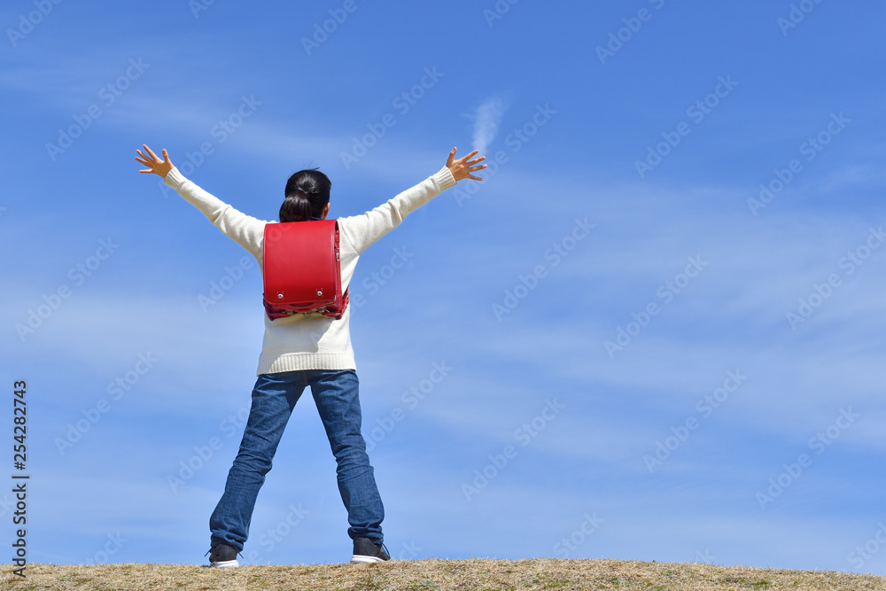 青空でバンザイする小学生の女の子（後姿）