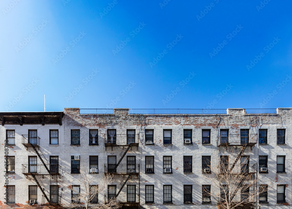 Old white brick apartment building with windows and fire escapes and an empty blue sky background overhead in New York City