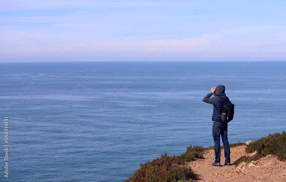 A man with a backpack looks through binoculars into the distance of the ocean. Cabo da Roca, Portugal. Cropped shot, horizontal, side view, perspective view. Concept of hobby and rest.
