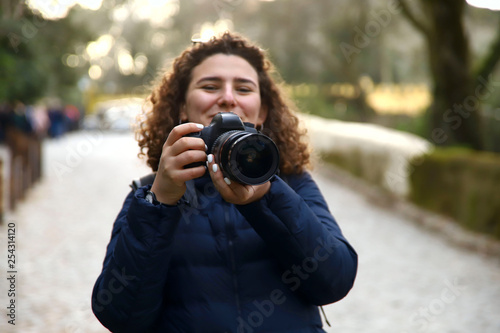 A smiling girl with curly hair holding a camera in the park on bokeh background. Cropped shot, horizontal, front view, perspective view. Hobie and leisure concept.