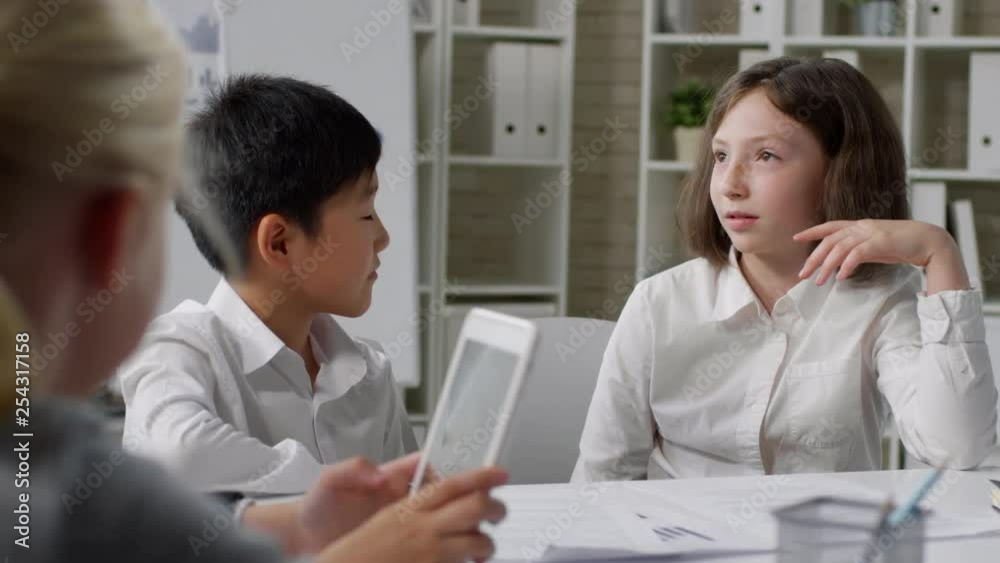 Medium shot of Asian boy and Caucasian girl sitting at desk and working in pair during lesson Medium shot of Asian boy and Caucasian girl sitting at desk and working in pair during lesson