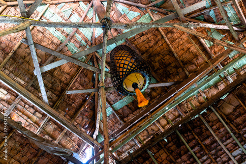 Interior of roof made of dried palm leaves and bamboo stick structure with hanging black-framed lamp made of bamboo and yellow pompom at a home stay in hill tribe village.