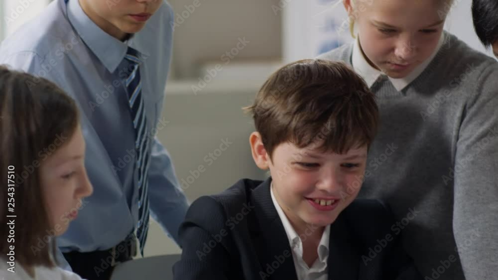 Tilt down of school kids surrounded Caucasian boy with tablet in his hands, watching pictures and smiling
