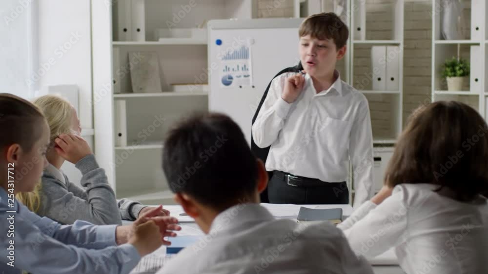 Panning of several kids sitting at desk and listening to their little business colleague walking from side to side, holding blazer and speaking