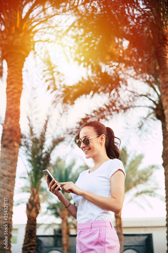 Under the sheen. A photo of a girl staying in the center of diversity of palm trees and yellow sun lights. She is wearing white and rose outfit with sunglasses  while using her phone.