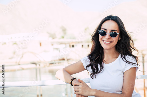 Blanc de chine. Faultless girl right in front of the camera in white t-shirt and black oval sunglasses with curly long brownish hair and sincere smile all over her hair in front of chalky buildings.