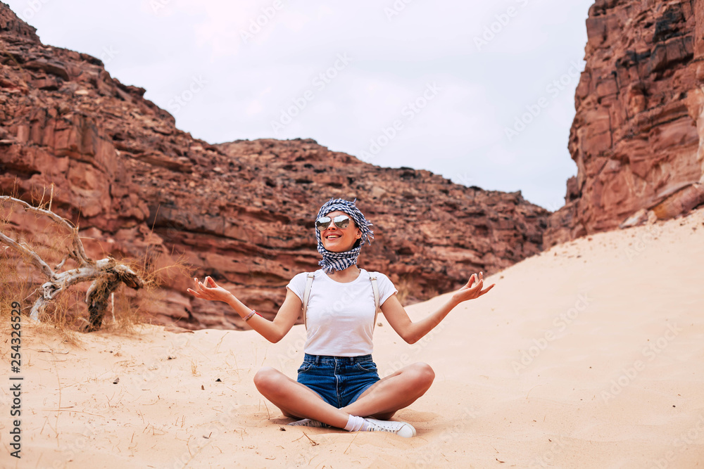 Woman in shorts, t-shirt, sneakers and kerchief is sitting on the soft sand and catching the peacefulness. The picture framed with the sky and brownish rocks.