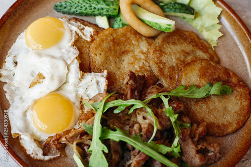 Delicious potato puncakes lying on brown clay plate with fried eggs. Fresh green cucumbers, fried golden onion and crusty meat, served with salad leaves. Tasty mouthwatering morning's meal. photo