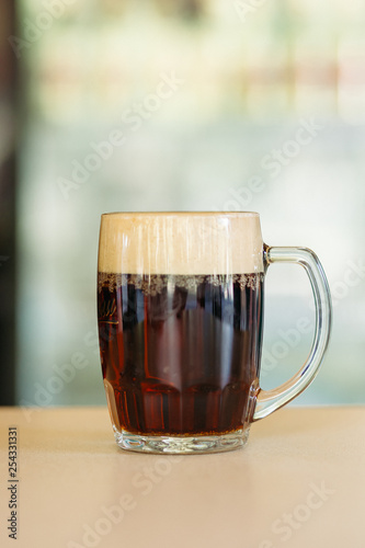 Photo of glass beer cup standing on smooth wooden surface. Cold dark summer drink for day heat. Fresh with thick high foam. Light blurred background. Concept of light alcohol drinks shooting.