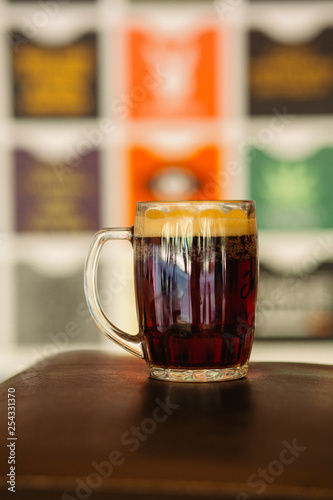 Photo of glass beer cup standing on smooth wooden surface. Cold dark summer drink for day heat. Fresh with thick high foam. Light blurred background. Concept of light alcohol drinks shooting.
