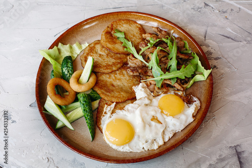 Delicious potato puncakes served with fried eggs laying on glancy clay plate including fresh green cucumbers, fried golden onion and crusty meat, served with salad leaves. Looking tasty. White table. photo