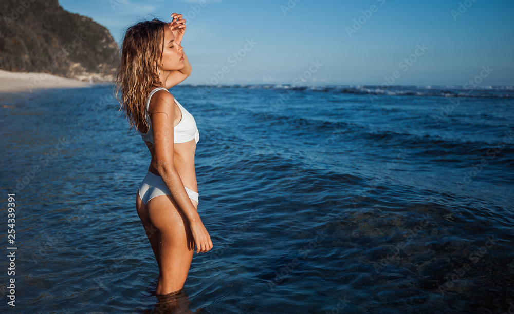 Girl in white swimsuit with beautiful tanned skin standing in the water on the beach Girl in white swimsuit with beautiful tanned skin standing in the water on the beach
