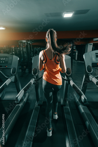 Slim brunette in orange sportswear and ponytail running on treadmill in gym at night. Backs turned. Do it now, sometimes later becomes never. © dusanpetkovic1