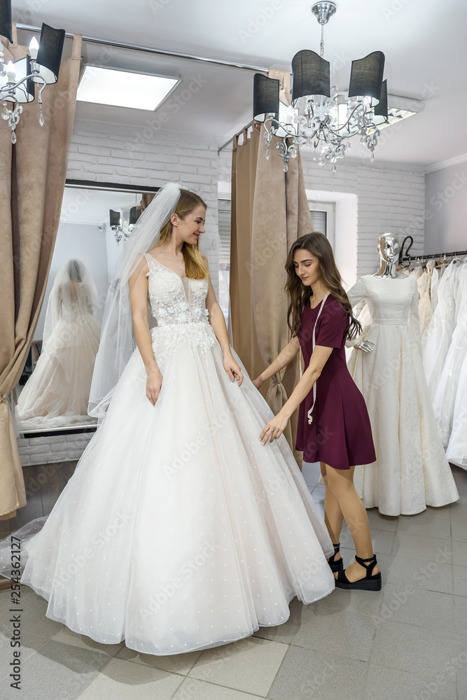 Tailor in wedding salon helping bride to try on  dress