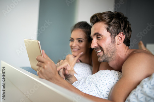 Happy young couple looking a tablet together and laughing while lying on the bed