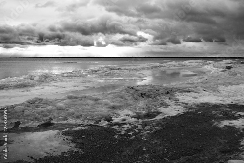 Lake Huron in the winter with snow and ice along beach with sand and rocks at sunrise in monochrome on a cloudy day with waves in the water. © zachary