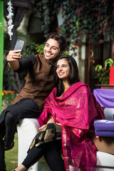 Smart indian couple taking selfie with smartphone on diwali or wedding party while sitting on sofa/couch