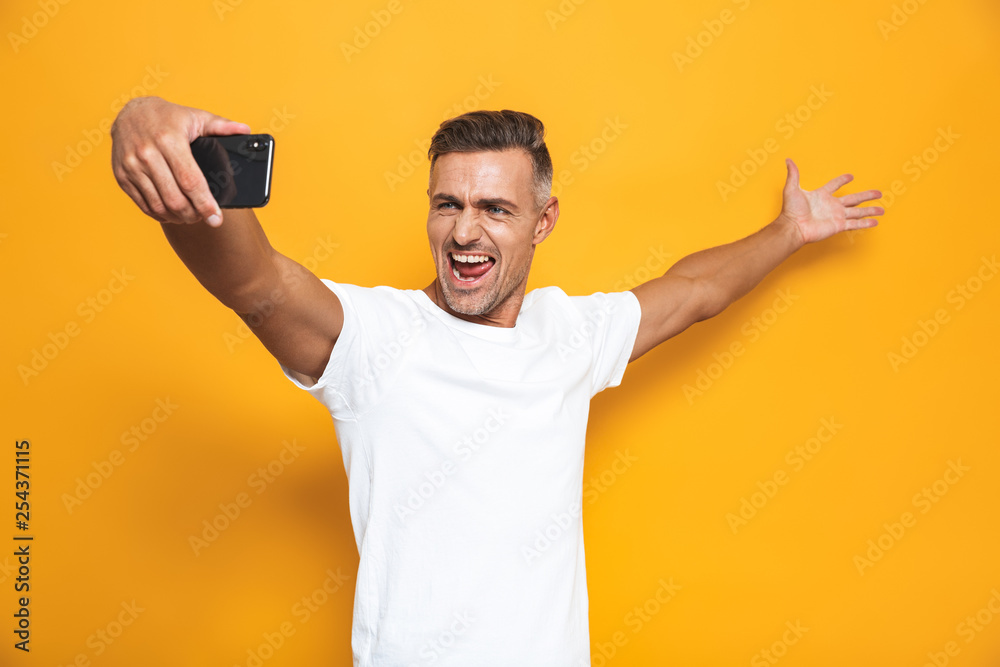Image of joyful man 30s in white t-shirt gesturing and taking selfie photo on mobile phone Image of joyful man 30s in white t-shirt gesturing and taking selfie photo on mobile phone
