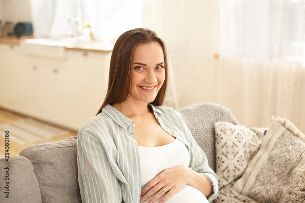 People, lifestyle, family and happiness concept. Indoor shot of cheerful attractive young pregnant female expecting her first baby, relaxing indoors on comfortable gray couch, smiling, touching belly People, lifestyle, family and happiness concept. Indoor shot of cheerful attractive young pregnant female expecting her first baby, relaxing indoors on comfortable gray couch, smiling, touching belly