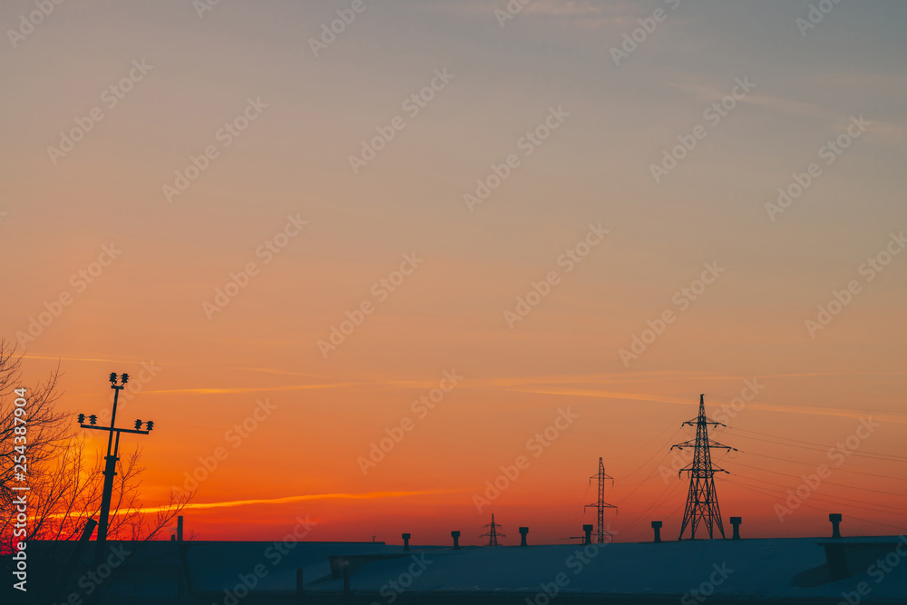 Power lines above roof on dawn. Silhouettes of tower with wires among smog on sunrise. High voltage cables on warm orange yellow sky. Power industry at sunset. City power supply. Mist urban background