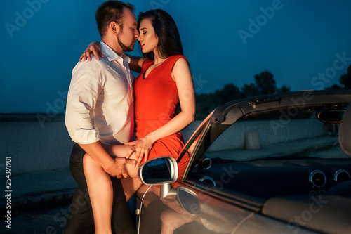 Gorgeous sexy copule of man and woman on a vacation car trip in summer day. Luxury grey sport car. Sexy fit brunette woman and man in evening outfit. Night portrait