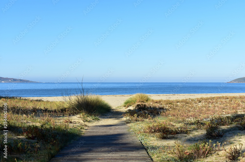 Beach with wooden boardwalk, grass, blue sea and clear sky. Galicia, Spain. Beach with wooden boardwalk, grass, blue sea and clear sky. Galicia, Spain.