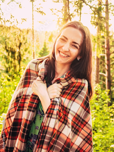Outdoors portrait of beautiful smiling brunette girl backlit. Beautiful caucasian brunette girl wrapped in a checkered plaid in the forest at sunset. Rays of the sun on the girl's face.
