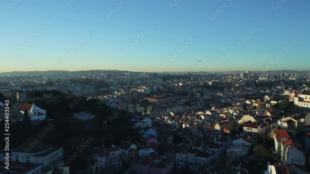 Aerial view; drone flight over the narrow streets of Portugal capital; panoramic landmark with last sunbeams and shade from the hill with Sao Jorge castel; aircraft flying in the background, Lisbon Aerial view; drone flight over the narrow streets of Portugal capital; panoramic landmark with last sunbeams and shade from the hill with Sao Jorge castel; aircraft flying in the background, Lisbon