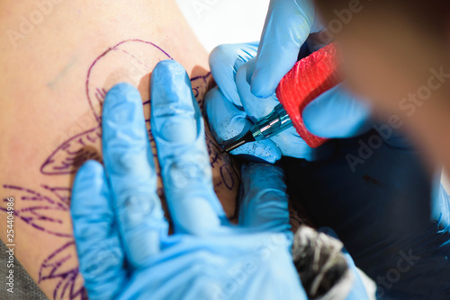 Master tattoo draws the black paint on the clients tattoo. Tattoo artist holding a tattoo machine in blue sterile gloves and working on the professional mat.