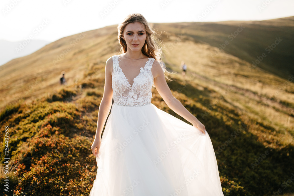 Stylish bride standing on beautiful landscape of mountains on sunset