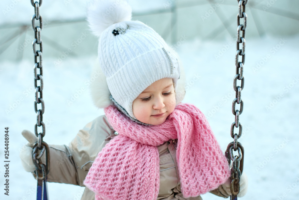 Happy child girl playing on a snowy winter walk. Little girl enjoys the game. Child girl playing on a swing outdoors in the snow. Street fun for the winter holidays. Happy child girl playing on a snowy winter walk. Little girl enjoys the game. Child girl playing on a swing outdoors in the snow. Street fun for the winter holidays.