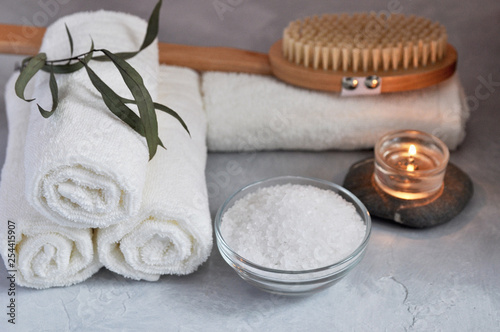 Close up view of Spa relax concept. White Terry towels, stones, candle, sea salt and wooden massage brush on a gray textured background.