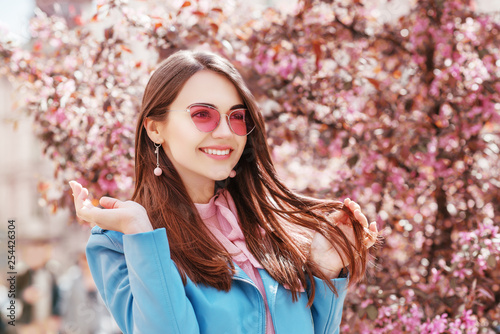 Outdoor close up portrait of  young happy smiling, laughing lady wearing stylish pink cat eye sunglasses, earrings, blue jacket, posing in spring street of European city. Copy, empty space for text