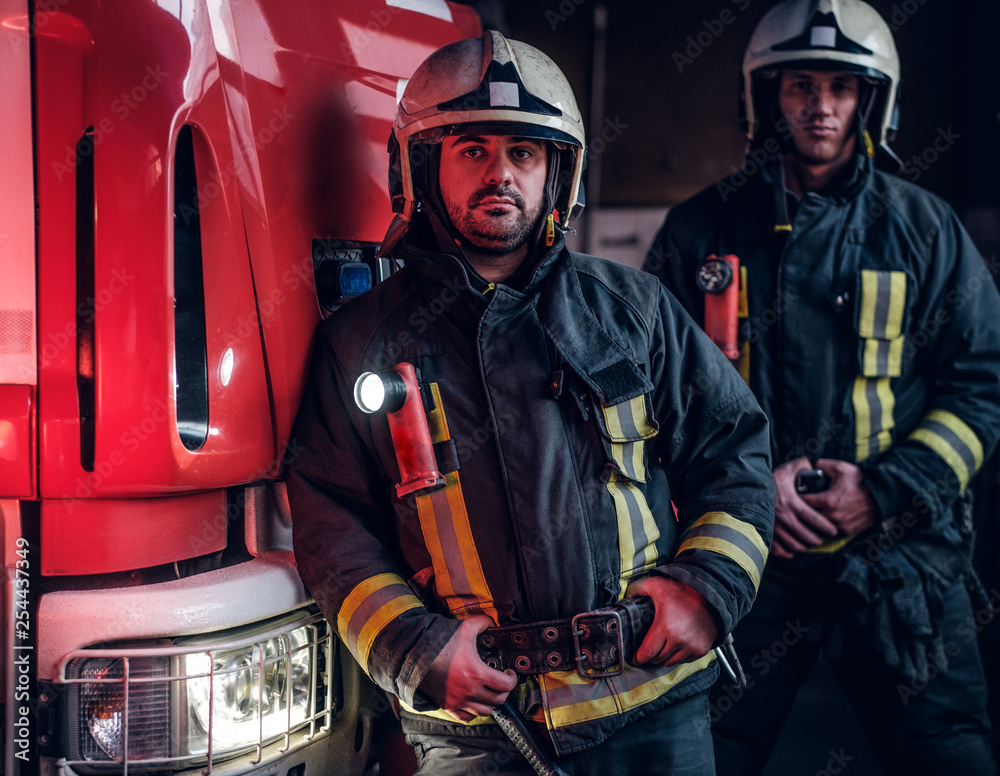 Obraz premium Two firemen wearing protective uniform standing next to a fire engine in a garage of a fire department.