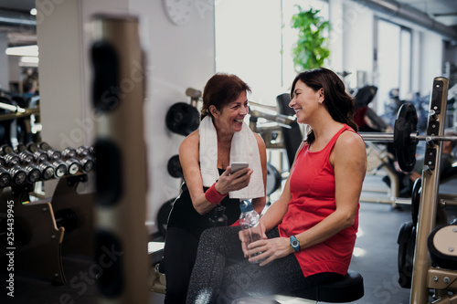 Two senior women in gym resting after doing exercise, holding smartphone.