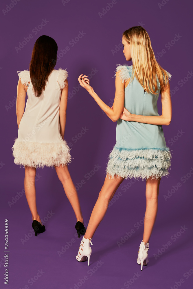 Professional photo of two young women, blonde and brunette, in cocktail dresses posing on purlpe background
