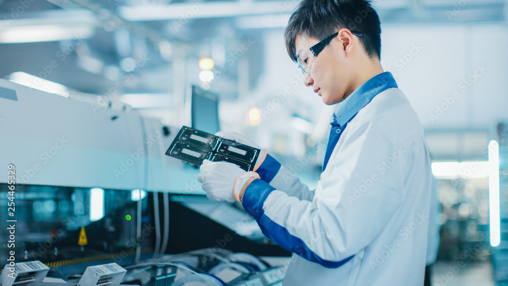 High-Tech Factory: Quality Control Engineer Checks Electronic Printed Circuit Board it for Damages. In the Background Assembly Line for PCB with Surface Mount Pick and Place Technology. High-Tech Factory: Quality Control Engineer Checks Electronic Printed Circuit Board it for Damages. In the Background Assembly Line for PCB with Surface Mount Pick and Place Technology.
