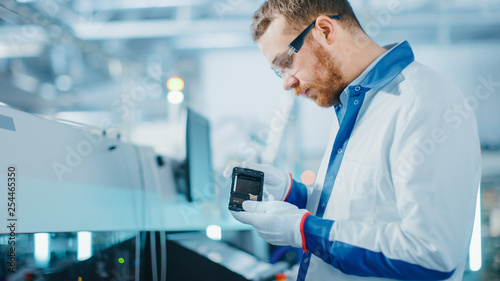 Canvas Print High-Tech Factory: Quality Control Engineer Picks up and Visually Checks Smart Electronic Device for Possible Damages