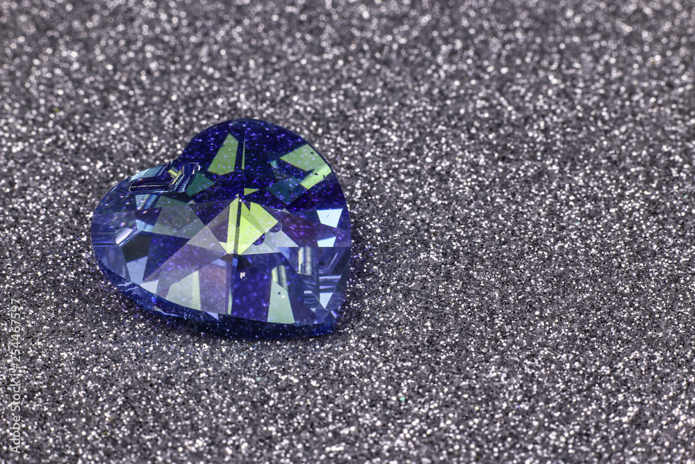 Blue heart-shaped facet crystal on the silver sparkling background. The light reflected in the precious stone facets Blue heart-shaped facet crystal on the silver sparkling background. The light reflected in the precious stone facets
