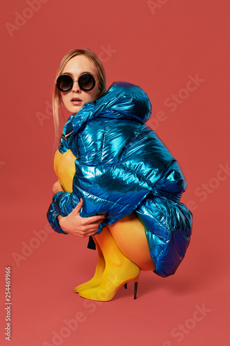 Young cute nice blonde girl in blue jacket sits in a fetal position in studio before camera, red background. Side view