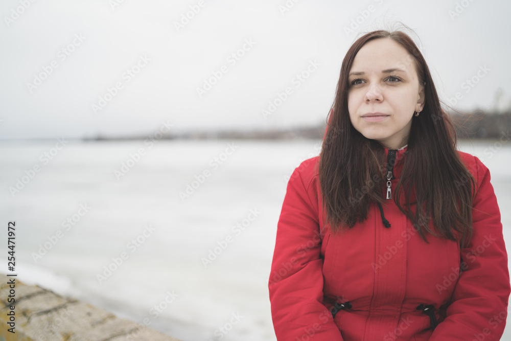 Beautiful girl in a red jacket on the pier in the spring posing against the water. Woman on the waterfront